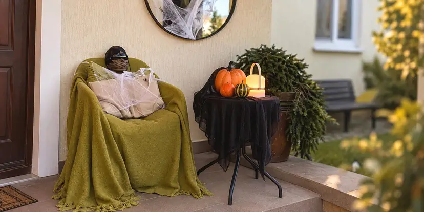 A Halloween porch: a 'mummy' sits in an armchair under a green throw, pumpkins and a lantern on a table with a black cloth, and a cobwebbed mirror above