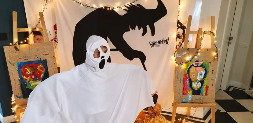 A person in a white ghost costume in front of a Halloween backdrop with fairy lights and children's paintings on easels