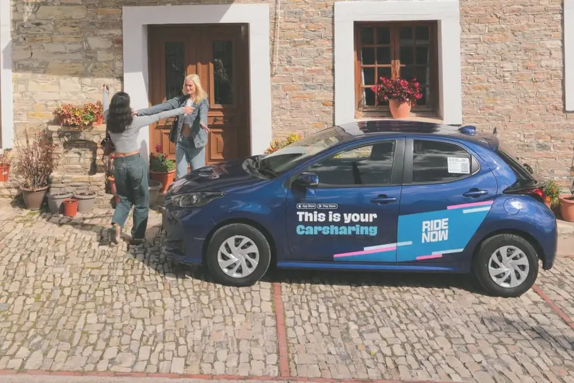 Ένα μπλε αυτοκίνητο της υπηρεσίας car sharing RideNow έξω από ένα πέτρινο σπίτι, με δύο νεαρές γυναίκες δίπλα του
